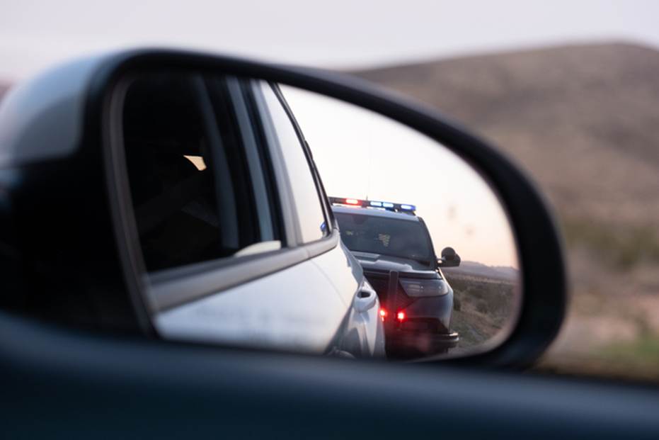 View of a police car with flashing lights in a car’s side mirror. Illustrates a topic of police misconduct, civil rights abuses and brutality cases.